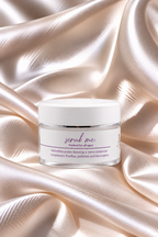 A clear jar of 'scrub me' facial scrub with a white lid and white label, set against a background of draped champagne-colored satin fabric.