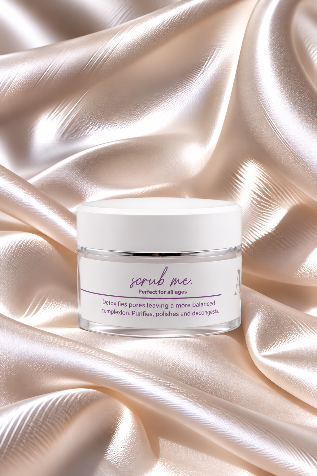 A clear jar of 'scrub me' facial scrub with a white lid and white label, set against a background of draped champagne-colored satin fabric.